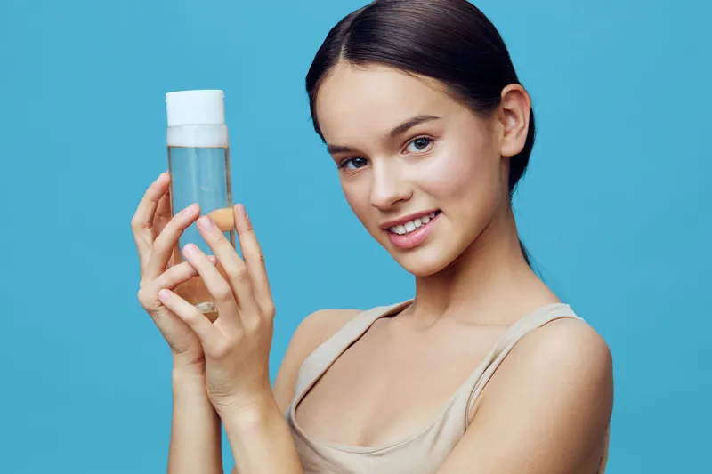 Young woman with a clear facial tonic on a blue background, smiling, showcasing healthy skin care routine for beauty and wellness