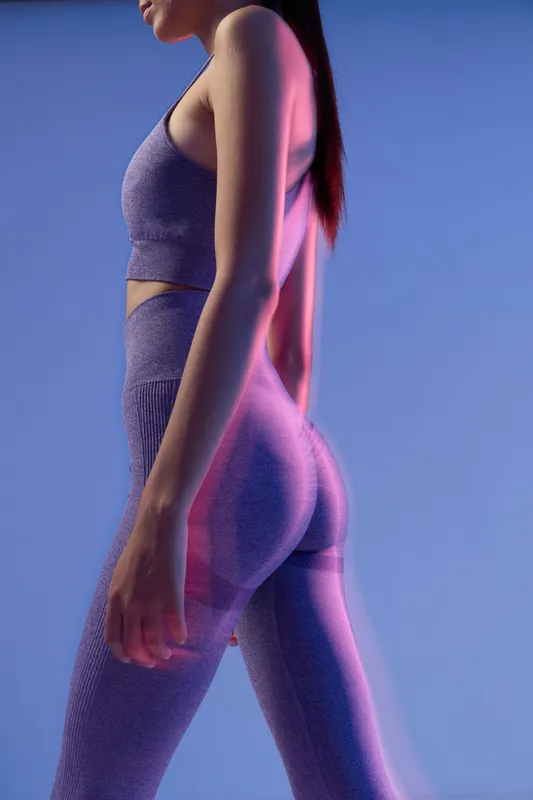 woman sport fitness wearing purple workout set poses in studio lighting, showcasing toned physique and active lifestyle with colorful neon shadows