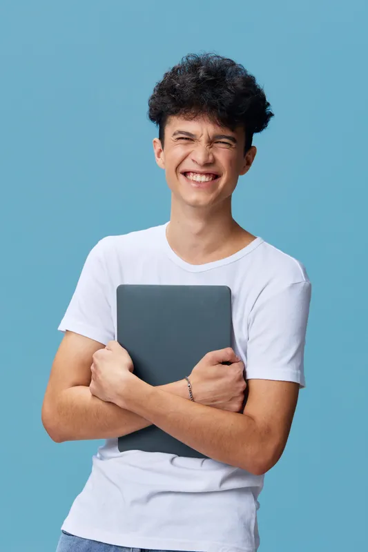 smiling man holding laptop with arms crossed, wearing casual white t-shirt, curly hair, blue background, cheerful young adult, technology concept