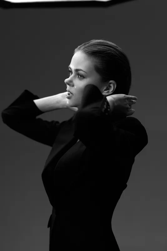 Black and white portrait of a woman adjusting her hair in a moody studio setting with dramatic lighting and stylish clothing.