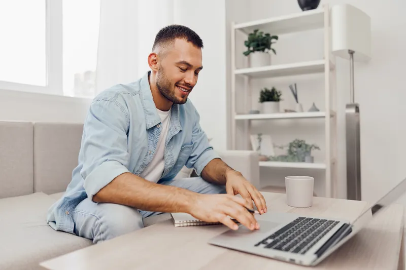 Smiling man working on laptop from home, reflecting modern remote work culture and embracing technology in a cozy environment. Focus on productivity and comfort.