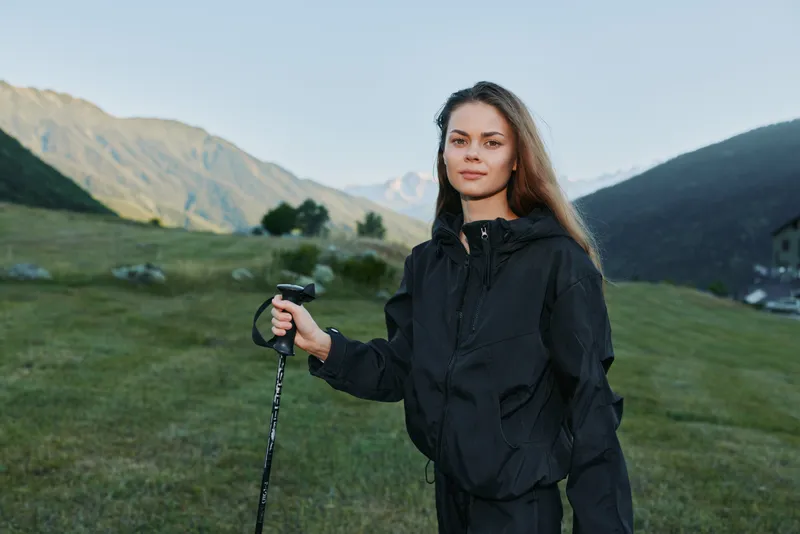 A confident hiker stands in a grassy mountain meadow, wearing a dark jacket and gripping a trekking pole. She looks ahead with calm determination, enjoying expansive nature and fresh alpine air.