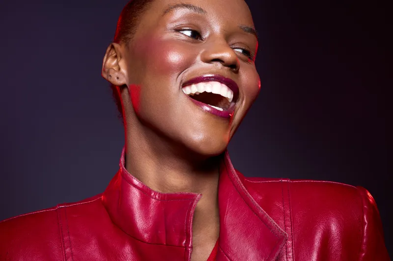 Happy African woman with dark skin laughing in red leather jacket. Studio portrait with vibrant makeup on purple background. Concept of joy, fashion, and confident beauty.