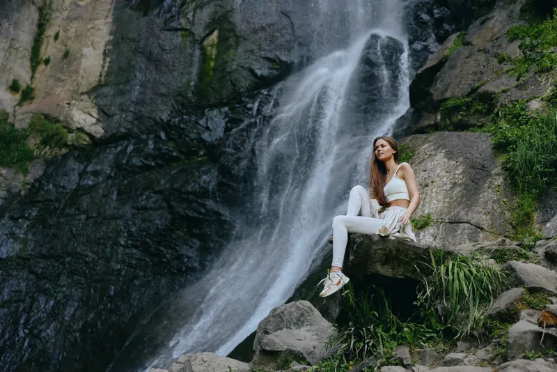 Young woman in white activewear sitting on a rock near a flowing waterfall surrounded by lush greenery in an outdoor nature setting.