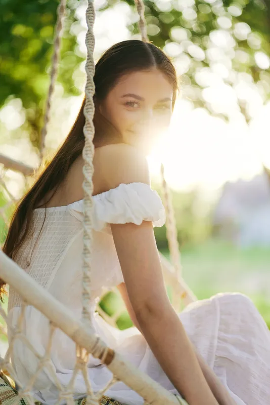 Beautiful young woman wearing a white dress, sitting on a swing, smiling in sunlight, surrounded by greenery, evoking a sense of joy and serenity