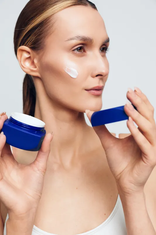 Young woman applying cream on face holding blue jar and lid close up, skincare routine, natural skin, beauty care, clean background, soft lighting