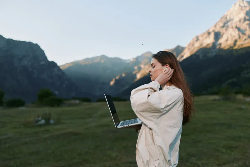 A cheerful woman stands outdoors in a scenic landscape with a laptop, wearing a light jacket. She smiles brightly, focused on work, surrounded by mountains and green fields.