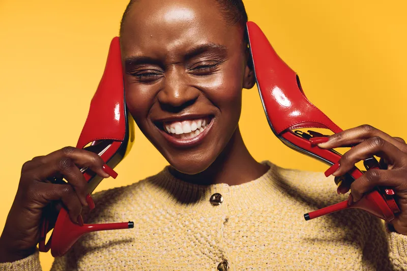 Happy dark skinned African woman smiles with closed eyes holding red high heel shoes near face. Studio portrait with warm lighting on brown background. Concept of fashion, joy, and beauty.