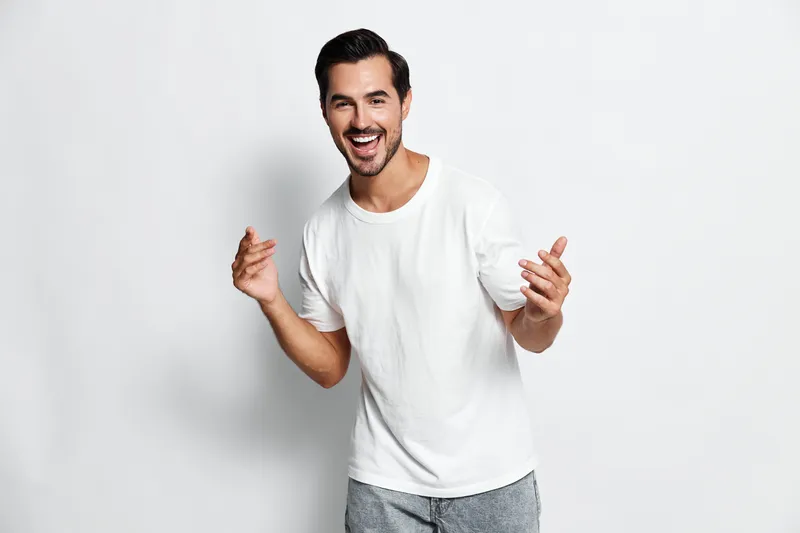 Full body smiling happy cheerful young man wear plain white t-shirt pose with hands lifted at camera against light neutral background. Joyful lifestyle concept.