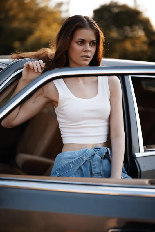 Young woman in a white crop top and denim jacket, posing in a vintage car during sunset, exuding a confident and carefree vibe.