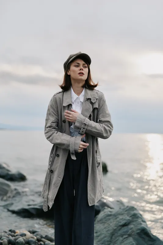 Young woman in grey coat and cap stands on rocky shore near calm sea at sunset. Soft natural light creates a peaceful atmosphere in cool tones. Concept of autumn fashion and solitude.