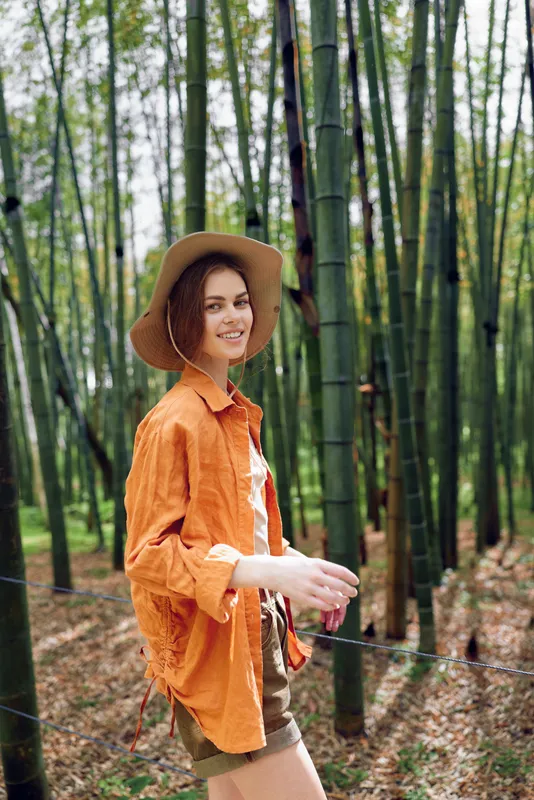 Woman in a hat and orange shirt walking through a bamboo forest, smiling at camera. Young female portrait in nature, casual summer outfit, sunlight, green grove and relaxed travel vibe.