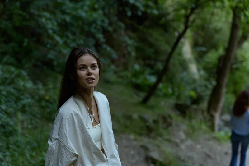 young woman in white jacket standing outdoors in lush green forest, natural light highlighting calm expression and casual style