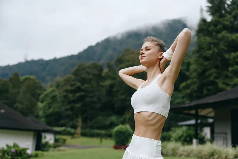 Athletic woman stretching outdoors in a serene green landscape, wearing a white sports bra and shorts, expressing joy and vitality amid lush nature and mountains