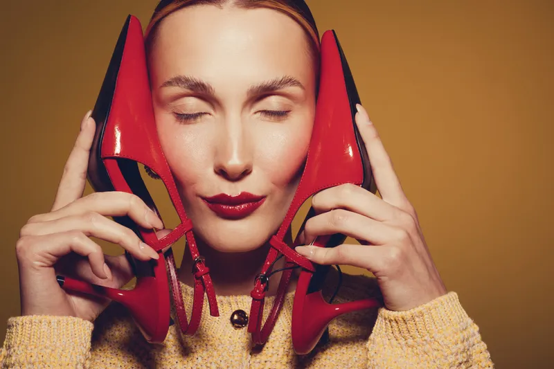 Woman with closed eyes holds red high heels near face, wearing yellow sweater. Concept of fashion, style, beauty, and footwear with warm studio lighting and expressive mood.