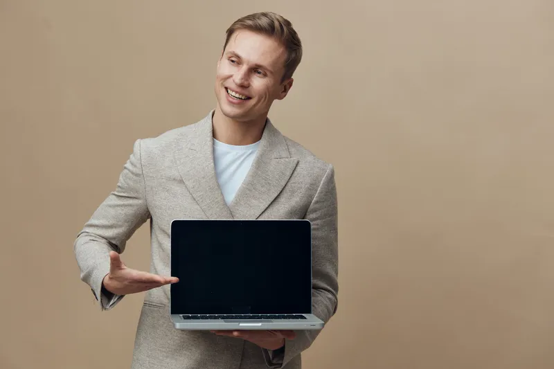 Confident young Caucasian man in smart casual blazer holding open laptop computer and pointing at screen isolated on beige background studio portrait. Business technology concept