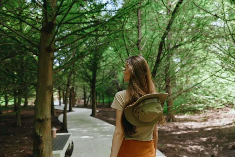 Woman walking on forest trail with backpack and hat, back view on shaded park path surrounded by green trees and nature, dappled sunlight and casual summer outdoor stroll.