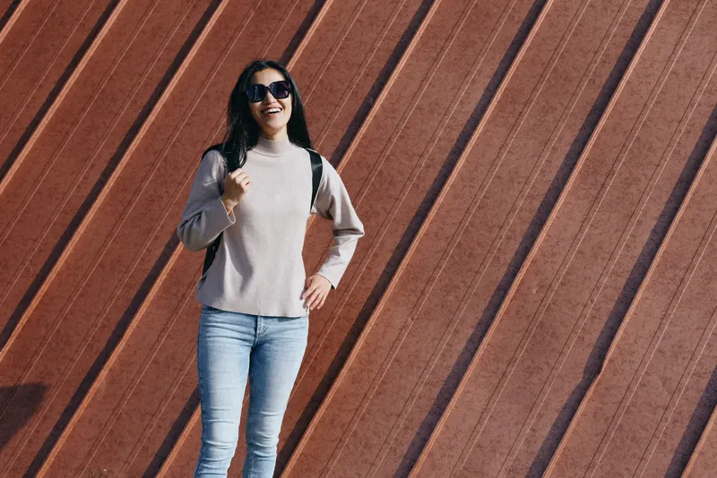 Woman wearing sunglasses and casual sweater poses against a diagonal wooden wall, outdoor fashion style, confident stance, urban backdrop and relaxed vibe.