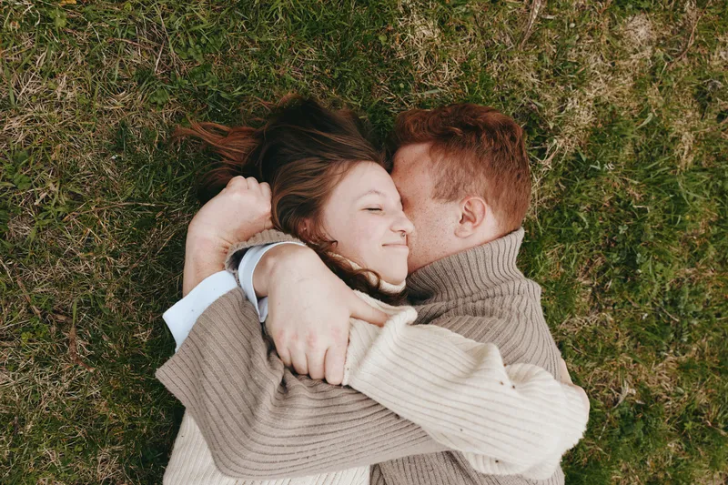 Couple lies on grass, embracing warmly, smiling and relaxed, wearing cozy sweaters, outdoor romance, intimate connection, peaceful mood, candid lifestyle moment under natural light, close together on