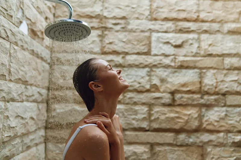 Young woman enjoying outdoor shower under warm water with natural stone wall background, smiling peacefully and feeling relaxed in bright daylight.