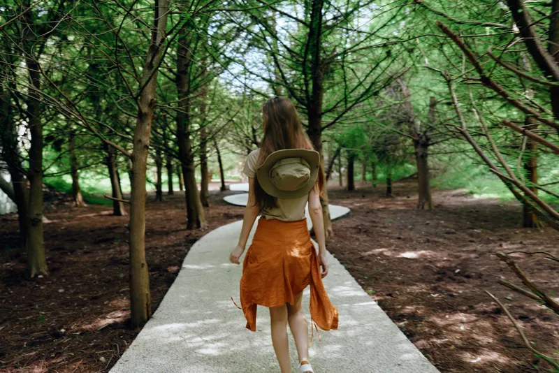 Woman with backpack and hat walking on a forest path and trail, rear view of traveler exploring nature and woodland, casual summer outfit on a paved walkway through trees.
