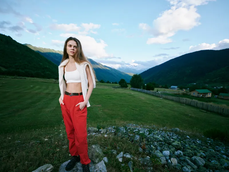 Woman outdoors on a grassy field with mountains in the background, wearing red pants and white top with a vest, casual fashion amid a scenic landscape and blue sky