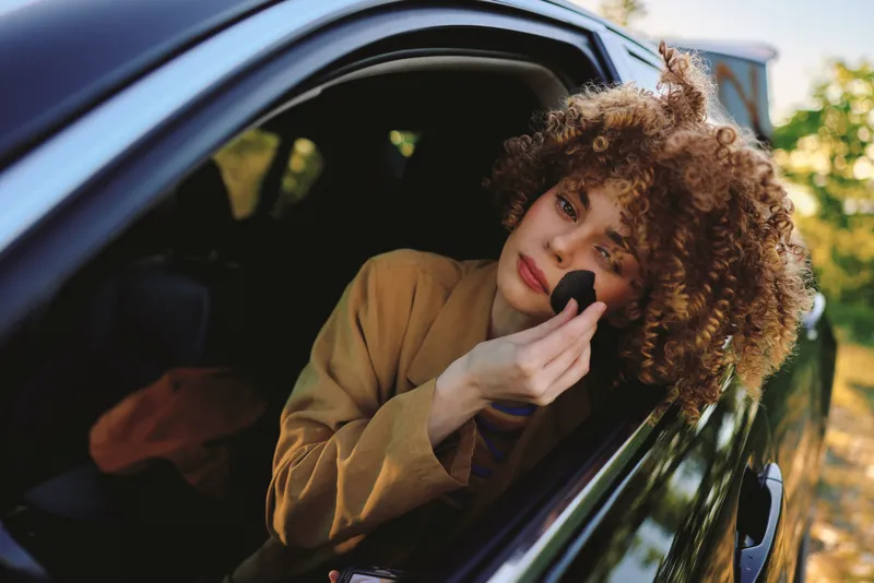 Woman with curly hair applying makeup in a car window, retro film color, lifestyle scene focusing on self care, beauty, and street style in a candid moment