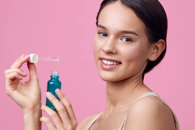 Young woman holding a skincare serum bottle, smiling joyfully, in a soft pink background, showcasing beauty and self care