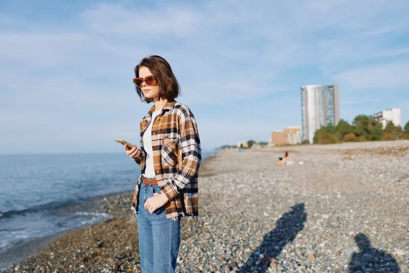 Woman on a pebble beach checks her phone, wearing a casual plaid shirt and jeans, sunglasses, with the sea, distant buildings and blue sky in the background.