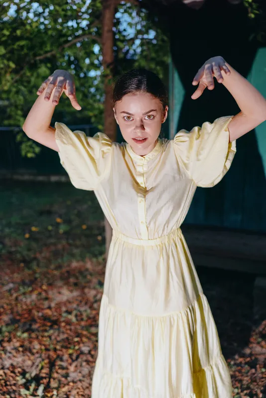Woman in a pale yellow dress performs a dramatic outdoor pose with raised arms among autumn trees and bright sunlight, creating a playful mood.