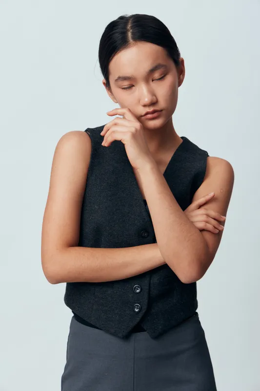 Young woman standing with crossed arms and eyes closed in thoughtful pose wearing dark vest and gray pants against plain light background. Portrait of calm or pensive female model.
