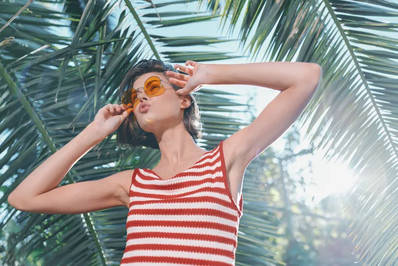 A joyful woman wearing a red and white striped tank top stands outdoors among tropical palm leaves, wearing sunglasses and smiling as bright sunlight glows, creating a vivid summer mood.