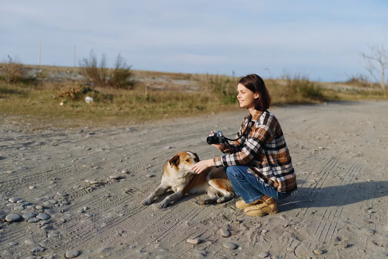 Woman photographer kneels on a rocky desert path, focusing on a small dog while holding a camera, capturing candid moments in an open sunlit wide landscape.