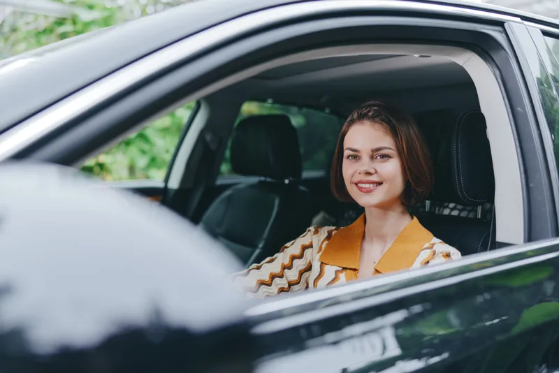 A cheerful woman sits in a car, smiling at the camera from the driver's seat, wearing a striped jacket and casual attire.