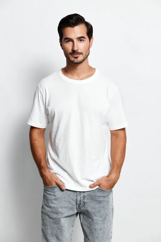 Confident smiling man in casual white t-shirt looks at the camera with hands in pockets, standing against a clean white background. Portrait of modern lifestyle.