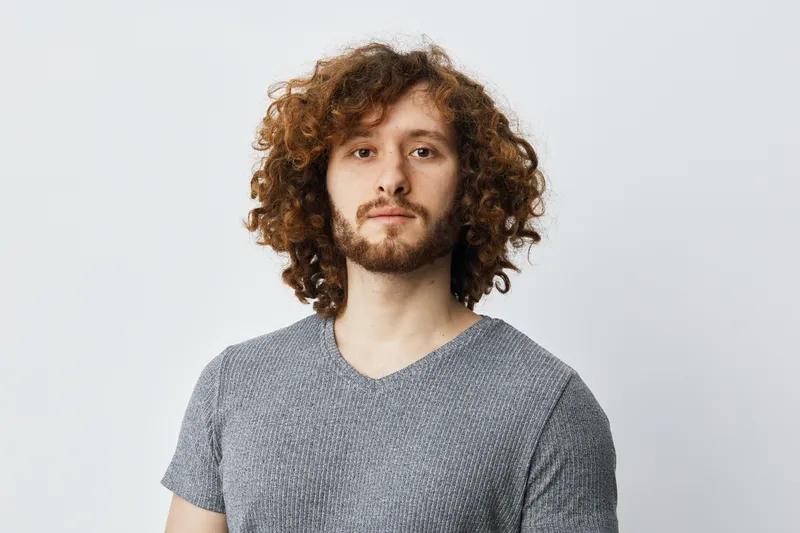 Portrait of a young man with curly hair, wearing a casual grey shirt, expressing confidence against a light background, suitable for diverse lifestyle themes