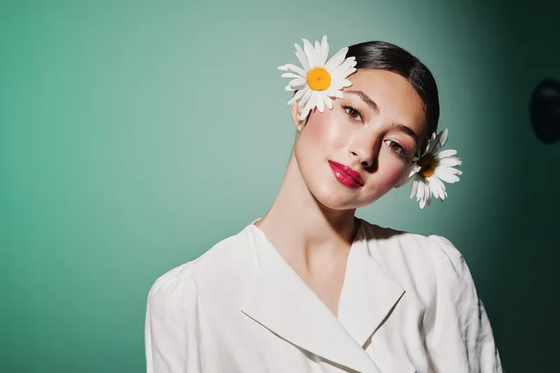Young woman with fresh natural makeup and vibrant red lipstick posing with white daisies in her hair against a pastel green background. Concept of beauty fashion creativity.