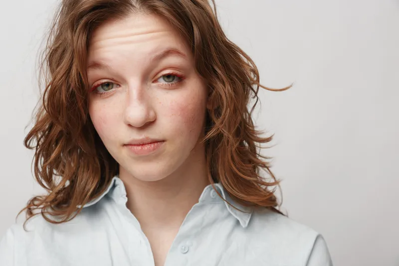 Young woman with curly hair and light freckles looks tired and skeptical wearing a white shirt on plain background. Concept for fatigue, doubt, and emotional expression.