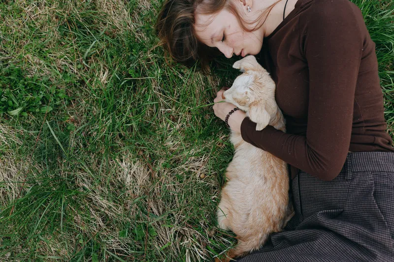 Carefree woman on grass gently cuddling a young goat kid, capturing a tranquil countryside moment. Natural light highlights affection, nature, and simple rural living.
