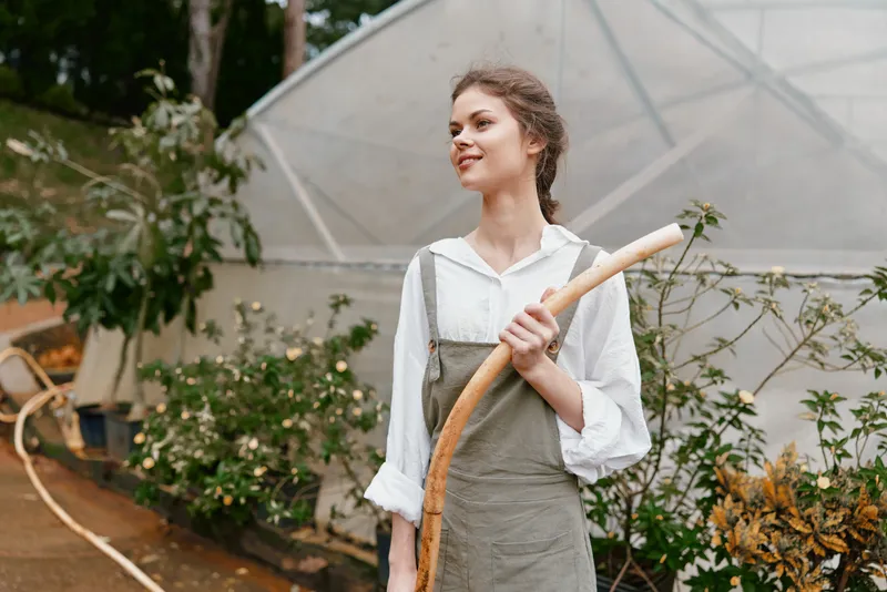 Young woman in casual gardening outfit smiles while holding a stick, with a greenhouse in the background, showcasing a vibrant nature concept