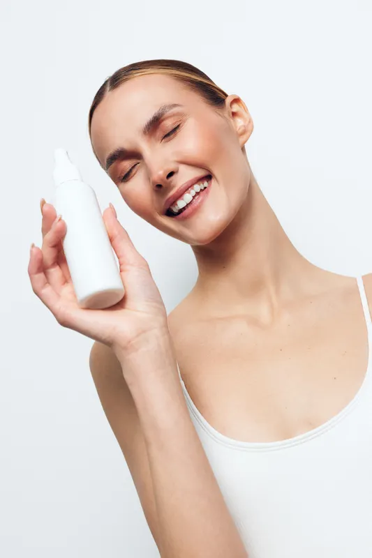 Woman smiling skincare face cream closeup selfcare wellness studio lighting, holding white bottle, applies moisturizer to face, clean light background, beauty routine