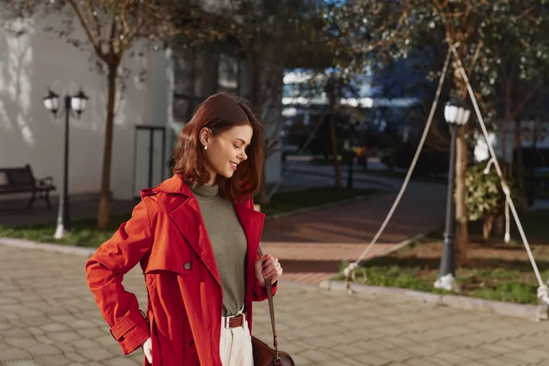 A young woman in a red coat smiles gently while standing outdoors on a sunny day with autumn trees and a bench in the background. Casual autumn fashion concept.
