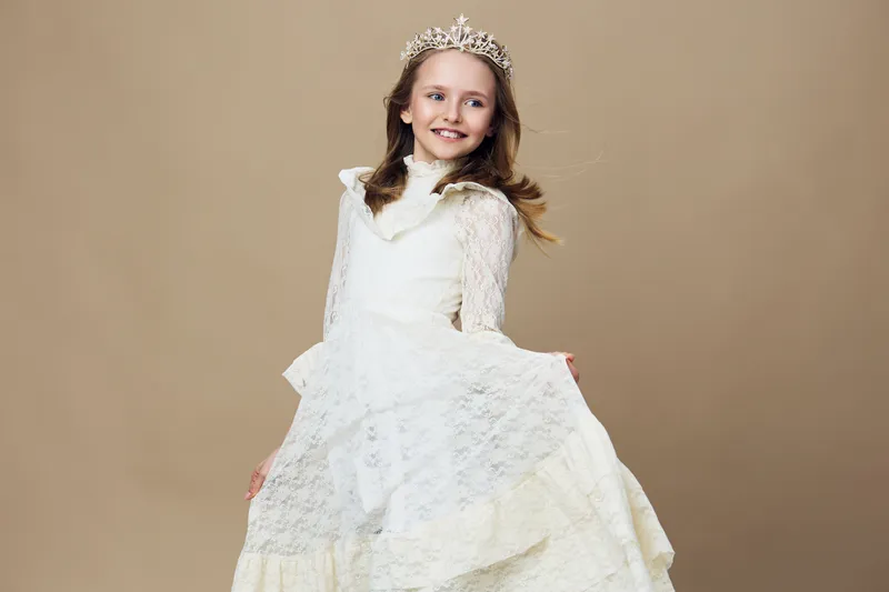 Happy girl in a white dress and crown, smiling on a neutral beige background, perfect for children s fashion and joyful moments