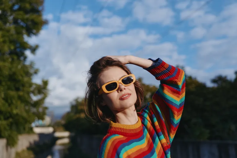 Woman in her 30s with short brown hair wearing yellow sunglasses and a rainbow striped sweater, smiling and posing outdoors by an urban park bridge for lifestyle fashion.