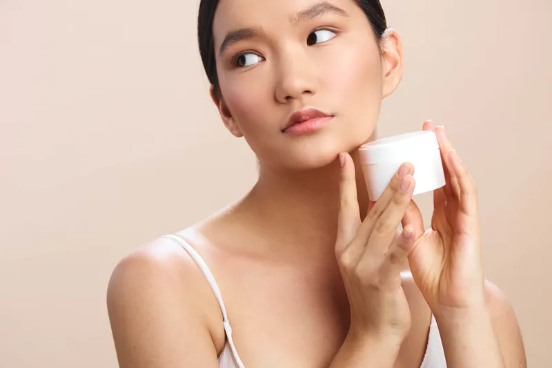 Asian woman applies moisturizer from a small jar in a clean, neutral setting. Calm expression and soft light convey personal care, wellness, and a positive skincare moment.