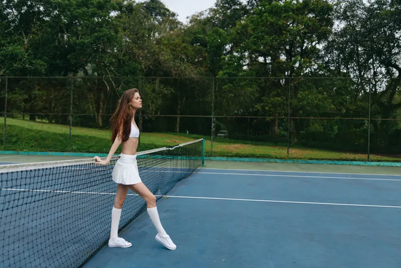 Young female tennis player in athletic wear poses confidently on a court, showcasing a vibrant daytime scene with greenery, reflecting energy, determination, and sportiness