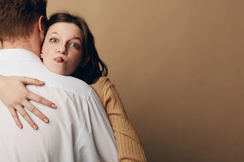 Young woman with brown hair makes funny face while hugging man in white shirt. Casual indoor moment with beige background. Concept of playful affection and relationship humor.
