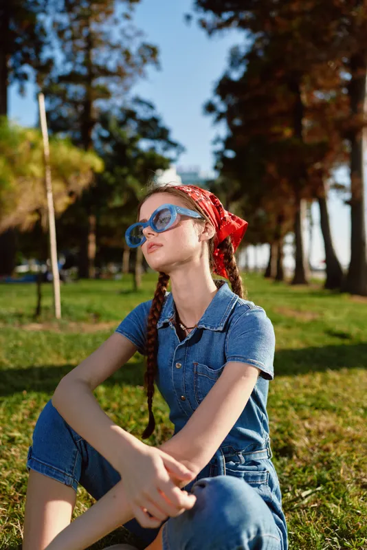 young woman sitting on grass outdoors wearing denim outfit, red headscarf and blue sunglasses enjoying sunny day in park