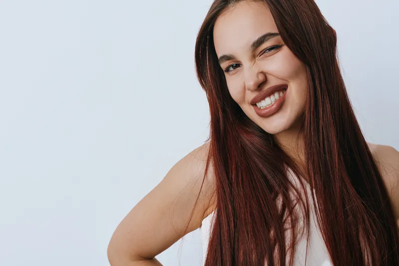 Young woman with long brown hair smiling and winking at camera on white background. Portrait of happy female with casual look and confident expression in studio light.