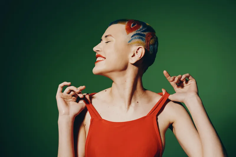 Young woman with colorful shaved hair and red dress smiling joyfully against a green background, expressing happiness and creative self-expression in vibrant style.
